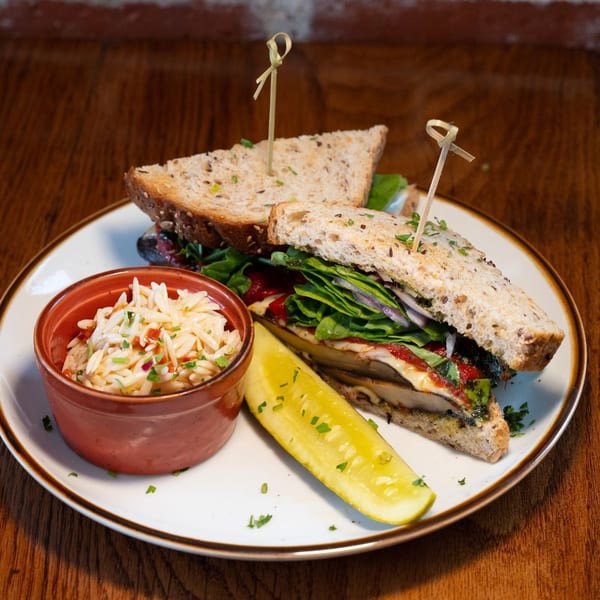 A grilled portobello sandwich with greens, red onion, and sauce, served with orzo salad and a pickle.