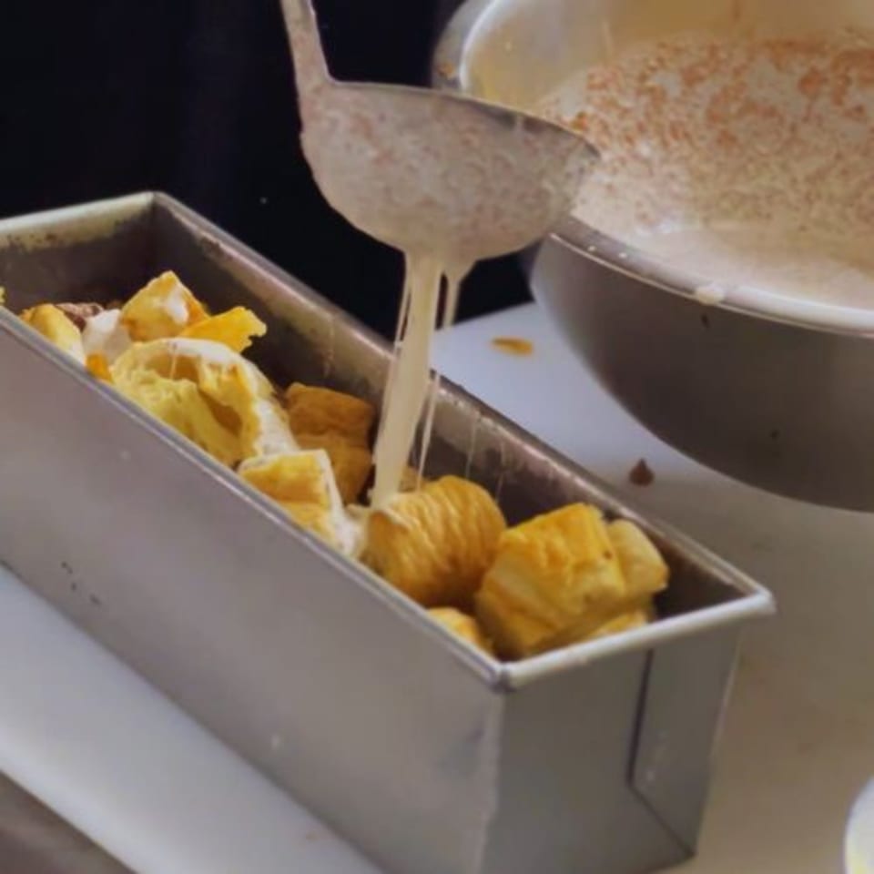 A ladle pouring a creamy sauce over golden bread pieces in a metal baking pan.