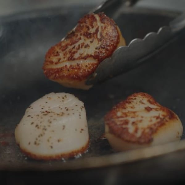 Three seared scallops in a pan, one being flipped with tongs, at Alyce cafe.