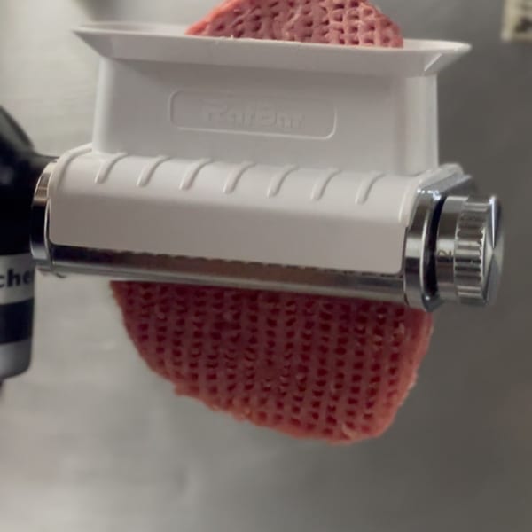 Close-up of a pasta maker extruding fresh, red pasta dough into a textured sheet.