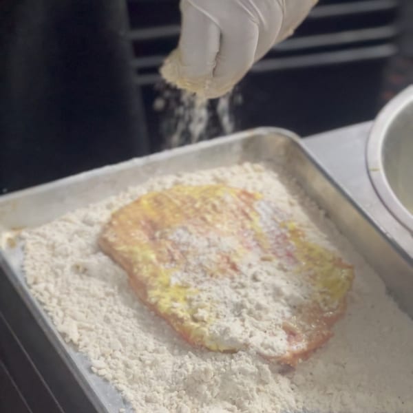 A gloved hand sprinkles flour over a piece of meat in a metal tray, preparing it for cooking.