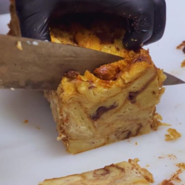 A gloved hand slices a large, golden-brown bread pudding with a knife on a white surface.