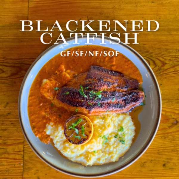 Blackened catfish with creamy grits, red sauce, and a grilled lemon slice on a wooden table.