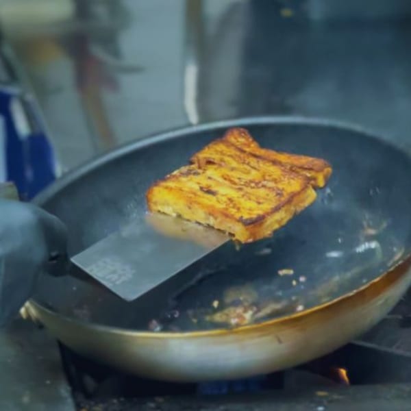 A golden-brown slice of French toast cooking in a pan, being flipped with a spatula.