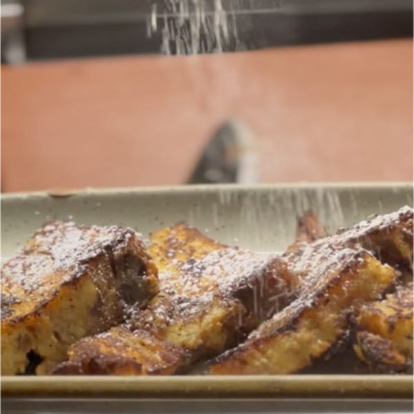 Powdered sugar being sprinkled over a plate of golden-brown French toast at Alyce cafe.